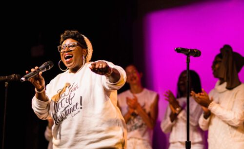 A Black woman from Ultraviolet performing on stage, looking happy while holding a microphone and wearing a sweatshirt with the words, 