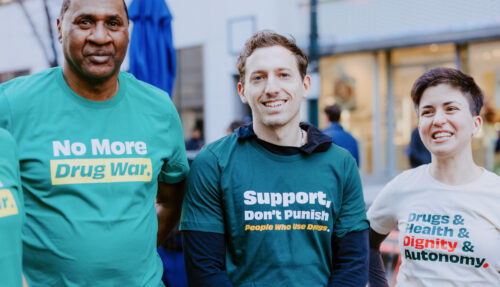 Three people standing together at a protest, wearing tshirts with positive slogans about drug policy and healthcare. The first says, 