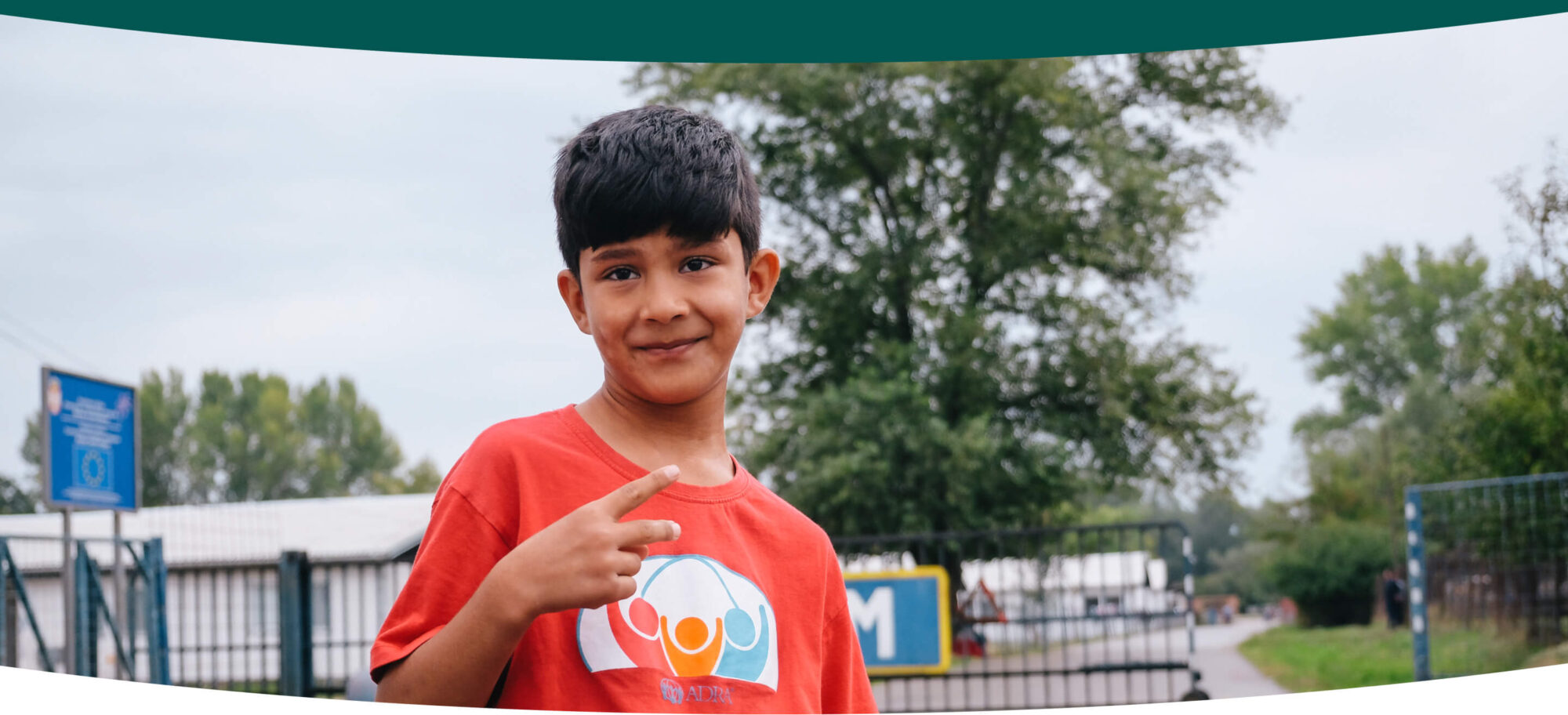 A young boy stands outside on a playground smiling and making a peace sign with his hand.