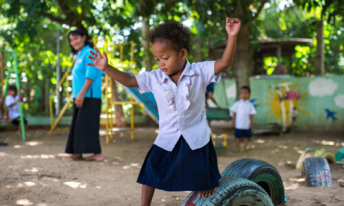 A young child in a school uniform balances on a tire in a playground while other children and an adult are in the background.