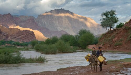 Boys on donkeys laden with water jugs walk along the edge of the Ajar river, as the surrounding red cliffs are lit by the setting sun, near Dehe Khankhala, upper Ajar Valley Bamiyan Province, Afghanistan