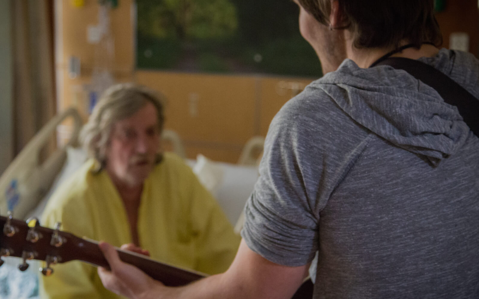 A young male musician plays guitar for an older man in a hospital bed.